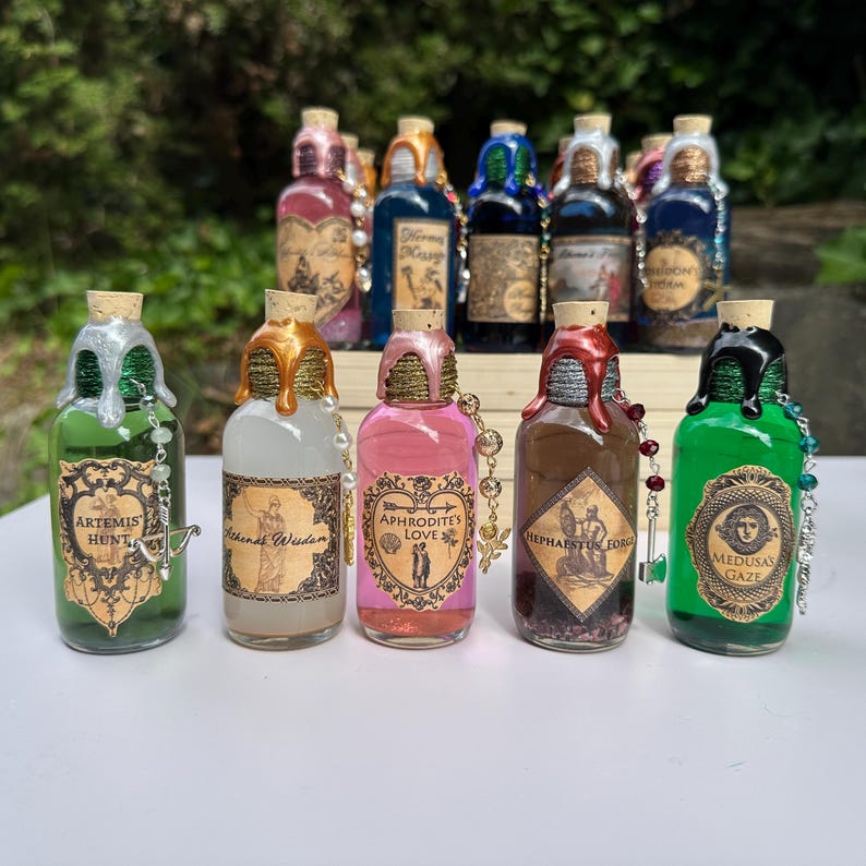 May include: A selection of decorative glass bottles with cork stoppers. Each bottle contains a different coloured liquid and is decorated with metallic drips and beaded chains. Labels feature various names and illustrations.