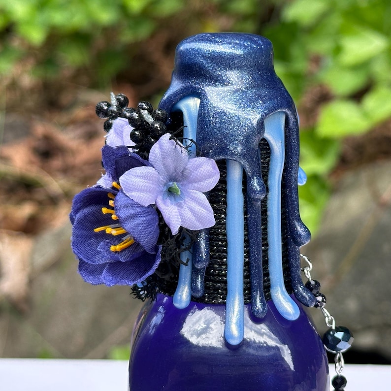 May include: A dark blue bottle with a decorative, glittery blue stopper. The stopper has a dripping design with light blue accents. A cluster of purple and black artificial flowers adorns the side, adding a touch of elegance.