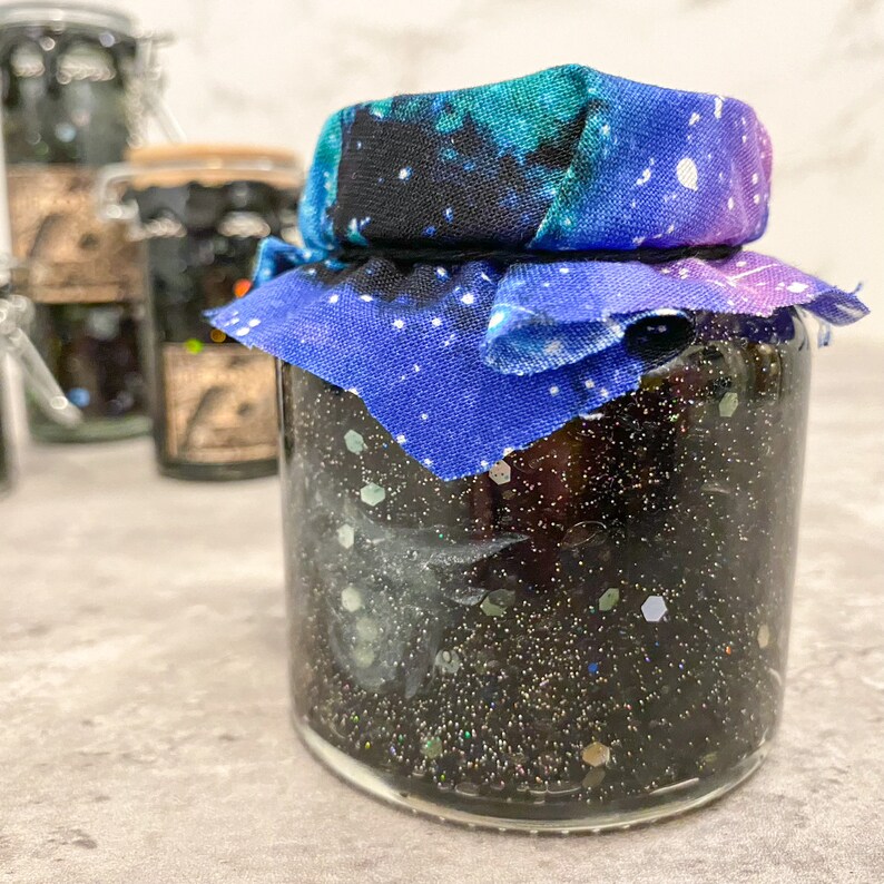May include: A glass jar filled with black glitter and a blue and purple galaxy print fabric tied around the lid.