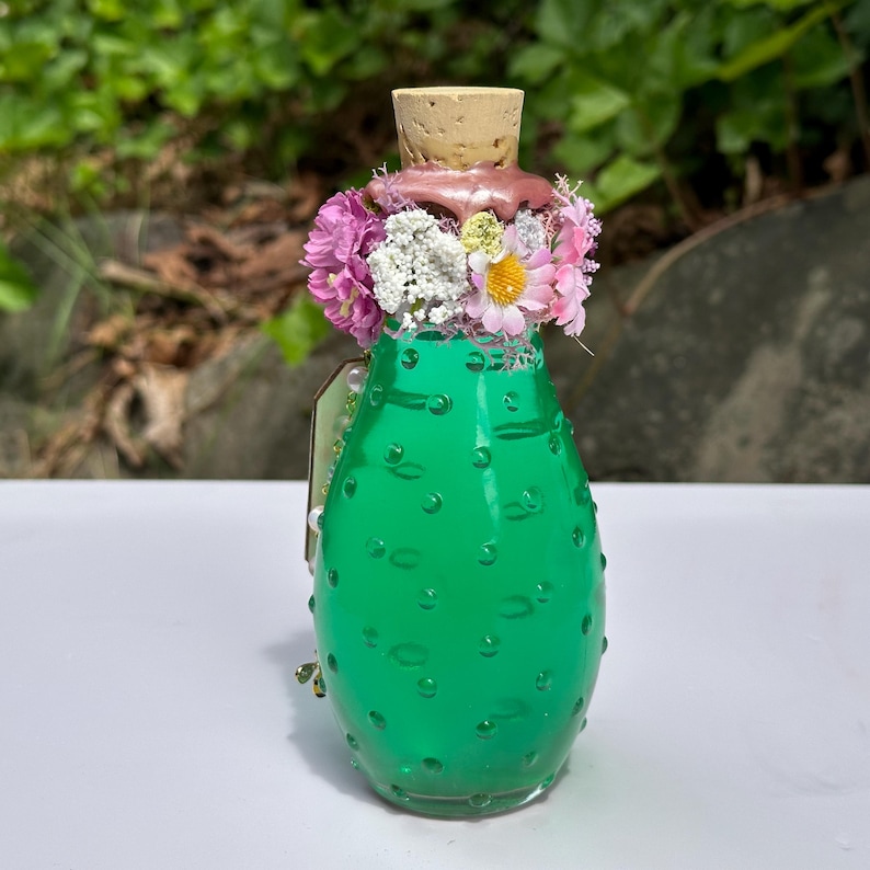 May include: A green glass bottle with a cork stopper, decorated with pink and white flowers. The bottle has a textured surface with small, round bumps. The top of the bottle is adorned with a pinkish-brown substance.