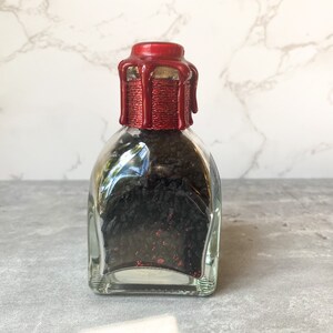 Puede incluir: Una botella de vidrio transparente con base cuadrada y un sello de cera roja en la parte superior. La botella est&aacute; llena de arena negra.