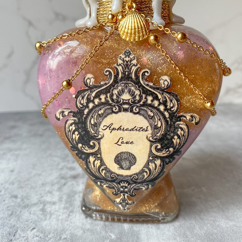 May include: A heart-shaped glass bottle with a gold glitter and pink liquid inside. The bottle has a gold chain and beads around the neck and a gold seashell charm. The bottle has a black and white label with the text "Aphrodite's Love" and a seashell design.