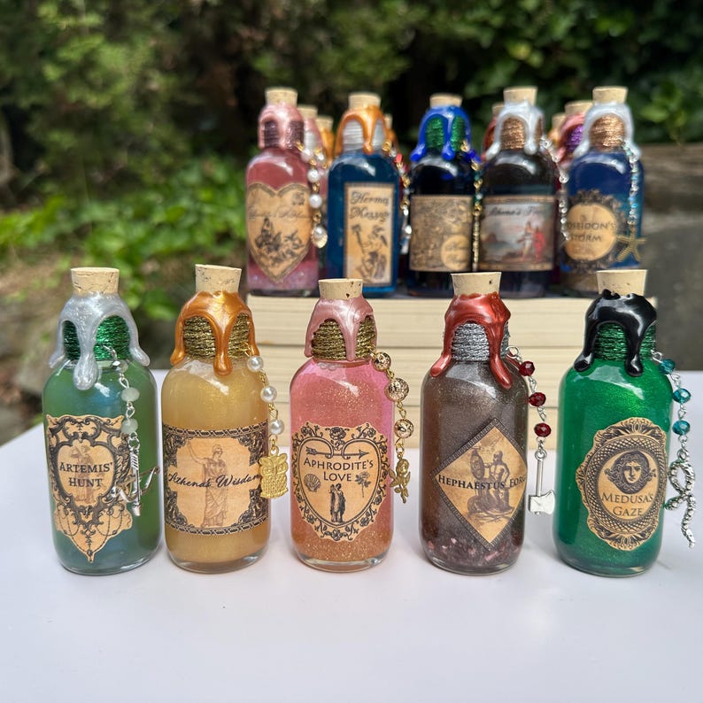 May include: A selection of decorative glass bottles with cork stoppers. Each bottle has a unique colour and label, with metallic accents and beaded embellishments. The labels feature text and illustrations.