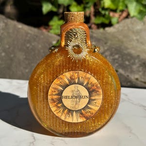 May include: A round, gold bottle with a cork stopper and a gold glitter finish. The bottle has a label with the text "HELIOS SUN" and a sun design.