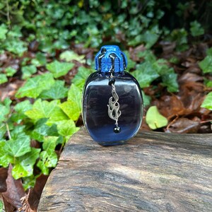 May include: A small, dark blue glass bottle with a black and blue dripping wax-like lid. A silver charm with a flame design hangs from the bottle.