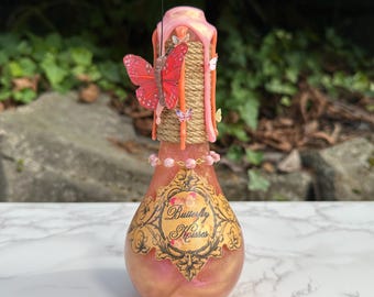 Peut inclure: Une bouteille décorative en verre contenant un liquide rose et doré. La bouteille est ornée d'une étiquette avec l'inscription "Butterfly Kisses". Le col est enveloppé de ficelle et surmonté de cire rose, d'un papillon rouge et de petits papillons décoratifs.