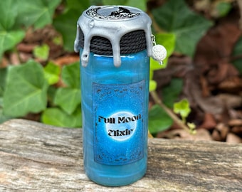 May include: A small glass bottle with a silver and black lid and a blue liquid inside. The bottle has a label that says "Full Moon Elixir" with a silver moon graphic.