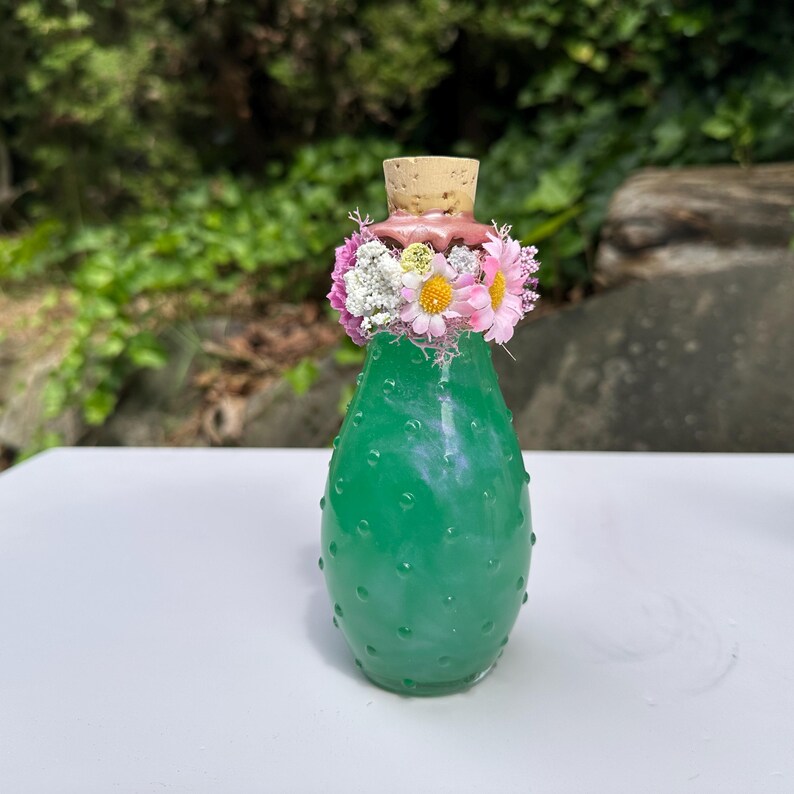 May include: A green glass bottle with a cork stopper, decorated with a pink wax seal and a floral arrangement of pink, white, and yellow flowers. The bottle has a textured surface. The background is a blurred green.