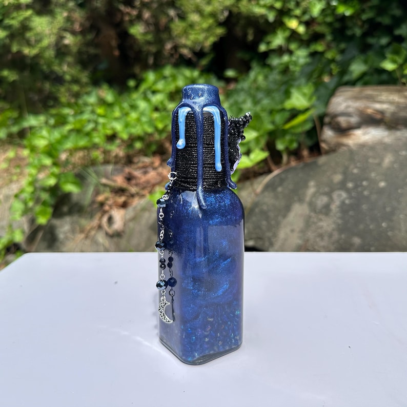 May include: A dark blue, square glass bottle with a decorative design. The bottle has a black and blue top with dripping blue accents. A beaded charm with a crescent moon hangs from the bottle.