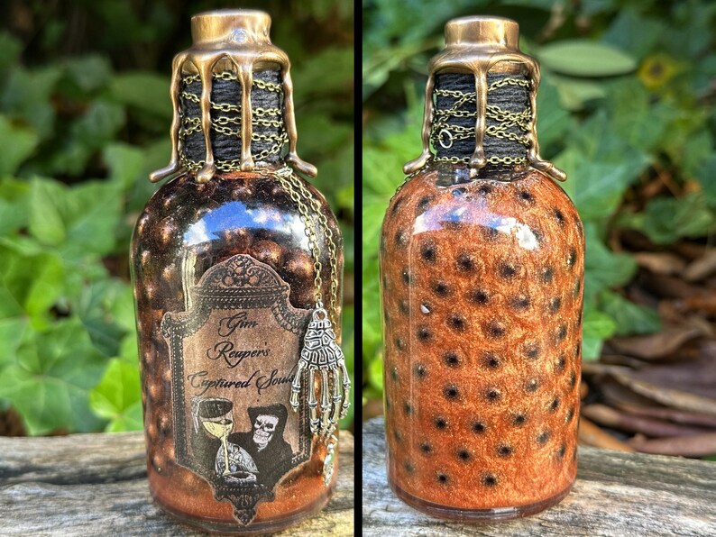 May include: A small glass bottle with a gold and black stopper and a label that reads "Grim Reaper's Captured Soul". The bottle is filled with a brown liquid and has a black and gold design.