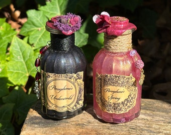 May include: Two small glass bottles with cork stoppers and decorative labels. The bottle on the left is black with glitter and says "Persephone's Underworld". The bottle on the right is pink with glitter and says "Persephone's Spring".