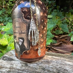 May include: A glass bottle with a brown and black label that reads "The Reapers Captured Souls". The bottle has a silver skeleton hand charm hanging from a chain.