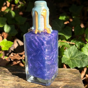 May include: A clear glass bottle with a purple liquid inside. The bottle has a gold and blue wax seal on the top.