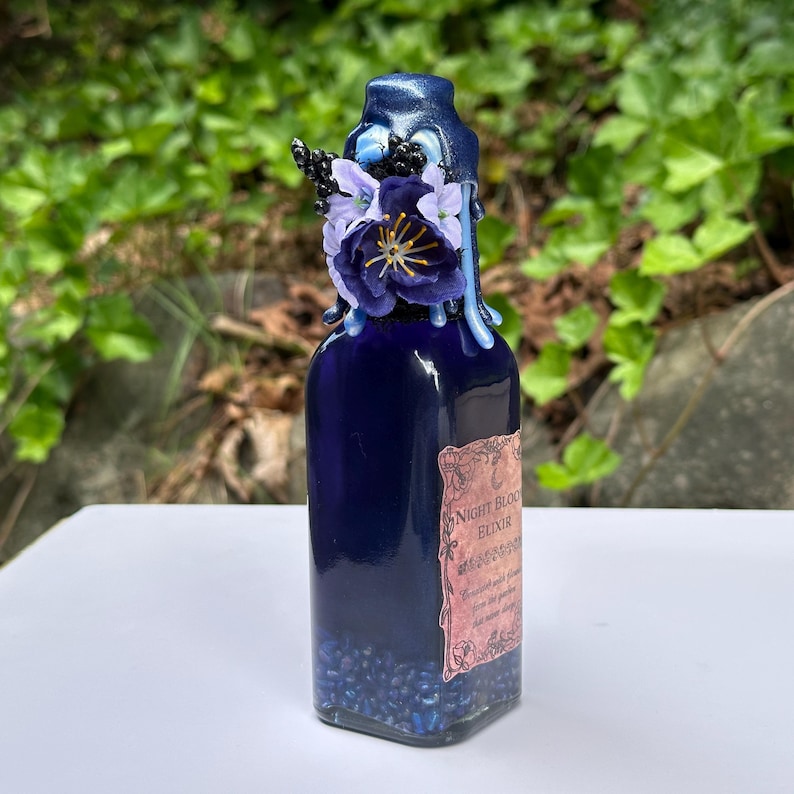 May include: A dark blue glass bottle with a decorative floral arrangement on top. The bottle has a label that reads "Night Bloom Elixir". The bottle is filled with small blue beads.