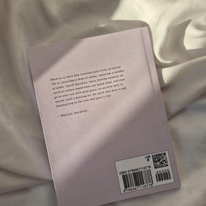 May include: A close-up of the back cover of a book with a quote by Marcus Aurelius. The book cover is a light purple color and the text is in black. The ISBN number and barcode are visible on the bottom right corner of the cover.