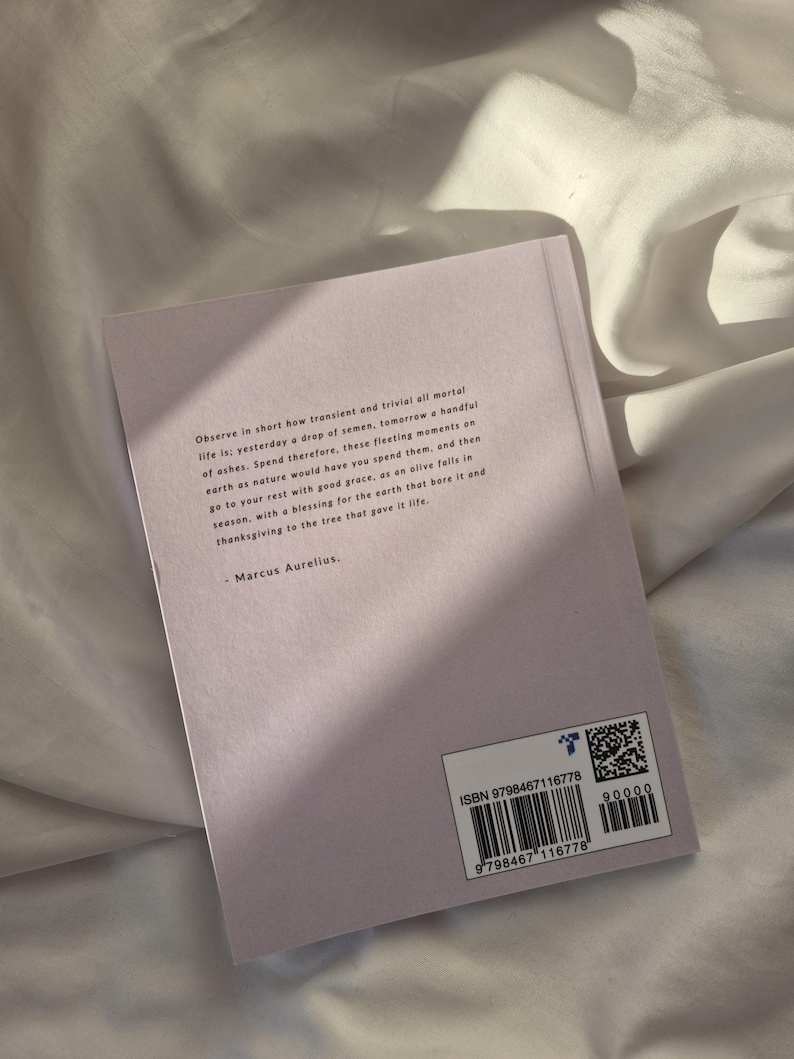 May include: A close-up of the back cover of a book with a quote by Marcus Aurelius. The book cover is a light purple color and the text is in black. The ISBN number and barcode are visible on the bottom right corner of the cover.
