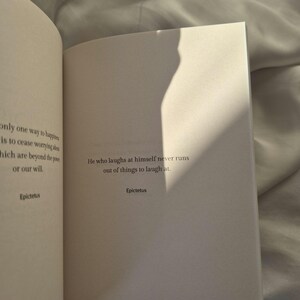 May include: An open book with a white background. The text on the page reads: "He who laughs at himself never runs out of things to laugh at." - Epictetus