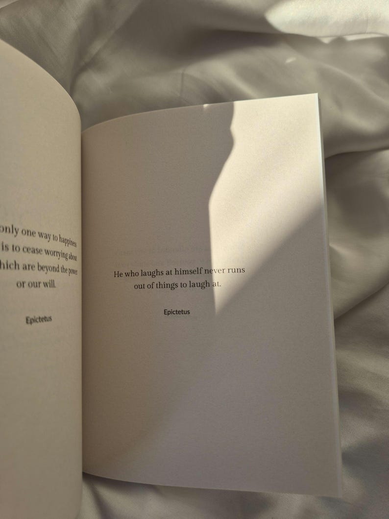 May include: An open book with a white background. The text on the page reads: "He who laughs at himself never runs out of things to laugh at." - Epictetus