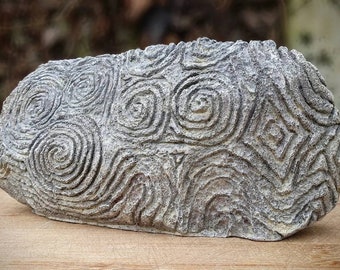 Puede incluir: Una piedra gris con un diseño tallado de patrones en espiral. La piedra es lisa y tiene una textura ligeramente rugosa.