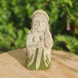 May include: A weathered stone statue of a figure with a crown and a sword. The statue has green moss accents and is set on a wooden surface against a blurred green background. The figure's face is detailed with carved features.