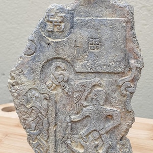 Könnte beinhalten: Eine verwitterte, graue Steinskulptur mit komplizierten Reliefdetails. Das Kunstwerk zeigt Figuren, Symbole und Muster, möglicherweise antiken Ursprungs. Der Stein ist grob rechteckig mit strukturierter Oberfläche.