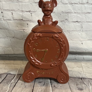 Puede incluir: Una jarra de galletas de cerámica marrón con forma de reloj con una figurita de ratón en la parte superior. La esfera del reloj tiene números y manecillas de color dorado. La jarra tiene las palabras "Time For Cookies" escritas en la parte delantera.