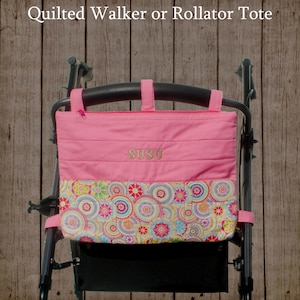 May include: Pink quilted walker or rollator tote bag with the name "SUSU" embroidered on the front. The bottom section features a colorful floral pattern. The tote has straps for attaching to a walker or rollator.