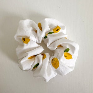 Op de afbeelding: Een witte scrunchie met een citroenprint. De citroenen zijn geel met zwarte pitten en groene bladeren.