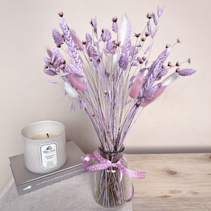 May include: A bouquet of dried lavender and white pampas grass in a clear glass vase with a pink polka dot ribbon. The vase is sitting on a wooden surface with a book and a candle.