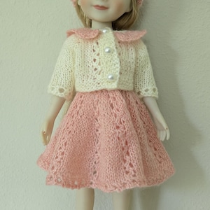 May include: A doll wearing a knitted outfit. The outfit includes a pink beret, a cream-colored jacket with pearl buttons, and a pink skirt. Matching pink shoes complete the ensemble. The doll has blonde hair and is smiling.