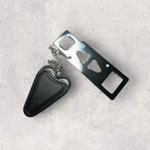 Könnte beinhalten: Ein silberfarbener Metall-Multitool-Flaschenöffner mit einem rechteckigen Körper und verschiedenen Aussparungen, der durch eine kurze silberne Kette mit einem schwarzen, dreieckigen Kunststoffteil mit glatter, matter Oberfläche verbunden ist.