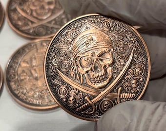 Puede incluir: Una moneda de color cobre con un detallado cráneo pirata que lleva un pañuelo y un parche en el ojo, espadas cruzadas y adornos ornamentados. La moneda tiene un diseño en relieve y una estética vintage.