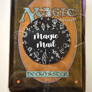 May include: A sealed deck of Magic: The Gathering trading cards featuring a brown and gold back design with the text Magic The Gathering and Deckmaster. A circular black sticker is centred on the plastic wrap with white line art and the text Magic Mail.