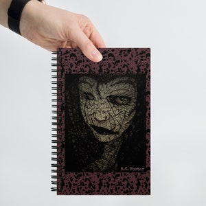 May include: A spiral bound notebook with a dark red cover featuring a black and white illustration of a woman's face with a textured background. The text "B.C. Foster" is printed in white at the bottom of the cover.