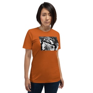 May include: A woman is wearing an orange t-shirt with a black and white abstract design. The design is a close-up of a swirling pattern.