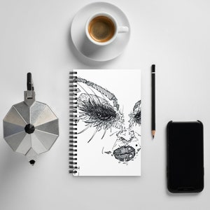 May include: A black and white line drawing of a woman's face with dramatic eyelashes and lips. The drawing is on a white spiral bound notebook.
