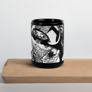 May include: Black ceramic mug with a white and black graphic design of a person with a large eye and a hand holding a small object. The design is intricate and detailed.