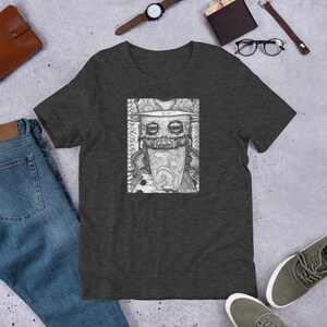 May include: A dark heather grey t-shirt with a black and white illustration of a creature with a hat and a large tongue. The creature has a creepy smile and is surrounded by a detailed pattern.