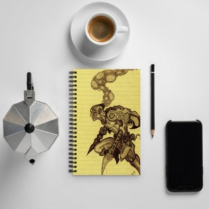 Può includere: Un quaderno a righe gialle con rilegatura a spirale. Il quaderno presenta un disegno a inchiostro nero e bianco di una creatura con fumo che esce dalla testa. Il disegno è su sfondo giallo.