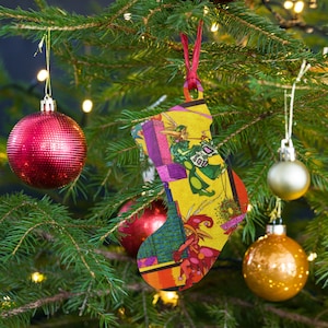 May include: A yellow Christmas stocking ornament with a colorful illustration of a whimsical scene featuring a jester, a fairy, and a cityscape. The stocking has a red ribbon and the text "Believe" is visible.
