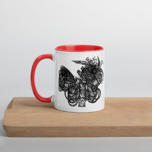 Puede incluir: Taza de cerámica blanca con borde rojo. La taza presenta una ilustración en blanco y negro de una persona con un brazo mecánico grande sosteniendo un arma.