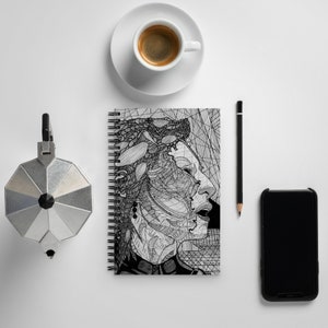 May include: Black and white line art illustration of a person's face on a spiral bound notebook. The notebook is closed and has a white background.