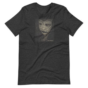 May include: Dark gray t-shirt featuring a detailed, stylized portrait of a woman with a cracked, textured appearance. The portrait is in a rectangular frame, with the text "B.C. Foster" below the image. The shirt is a crew neck.