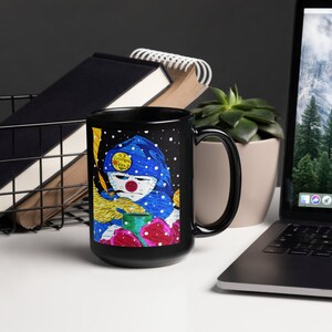 May include: Black ceramic mug featuring a colorful illustration of a figure with a blue hat, red nose, and a yellow object. The mug has the text "Is It Summer Yet?" on the hat. The design includes red and green floral elements.