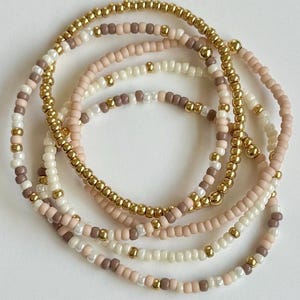 May include: A set of five stacked bracelets made from small round beads in metallic gold, matte beige, soft pink, and cream white. The beads are arranged in alternating patterns, creating a layered look with a mix of shiny and matte textures.