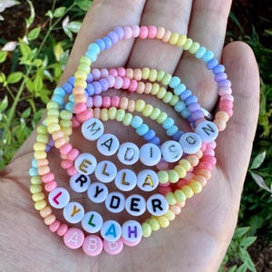 May include: A collection of colorful beaded bracelets in pastel shades of pink, yellow, blue, and purple. Some bracelets feature white letter beads spelling out names like "Madison", "Ella", "Ryder", and "Lylah". The bracelets are a fun and personalized accessory.