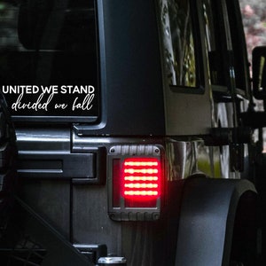 May include: A black Jeep with a white vinyl decal on the rear window that reads "United We Stand Divided We Fall". The Jeep has a red taillight and a black spare tyre.