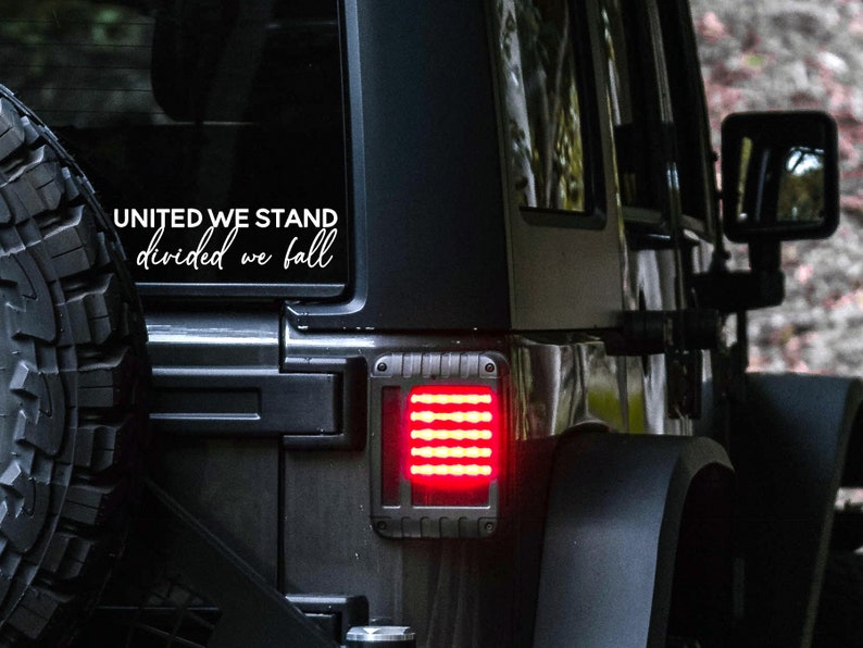 May include: A black Jeep with a white vinyl decal on the rear window that reads "United We Stand Divided We Fall". The Jeep has a red taillight and a black spare tyre.
