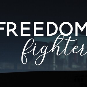Könnte beinhalten: Weißer Vinyl-Aufkleber mit dem Text "FREEDOM fighter" in einer kursivschriftlichen Schriftart. Der Aufkleber befindet sich auf einem dunklen Hintergrund.