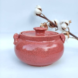 May include: A red ceramic pot with a lid and two handles. The pot has a speckled finish and is sitting on a white surface.